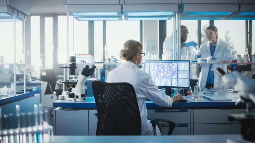 Medical Laboratory with Team of Scientists Working. Microbiologist is Using Personal Computer, Screen Shows High-Tech Concept for DNA research. High-Tech Biotechnology Research. Back View Shot Possible Benefits Of Participating In A Clinical Trial